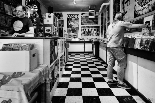 Black and white photo showing the interior of a record store. The floor is a checkerboard pattern and there's various posters on the wall. A guy is digging through the bins.