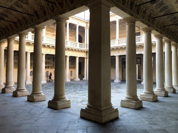 View of a pillared courtyard from one corner, with a square corner pillar in center of image and rounded pillars receding left and right.  Beyond them, light shines in from above to a two-story, very old courtyard.  A person can be seen walking on the far side.