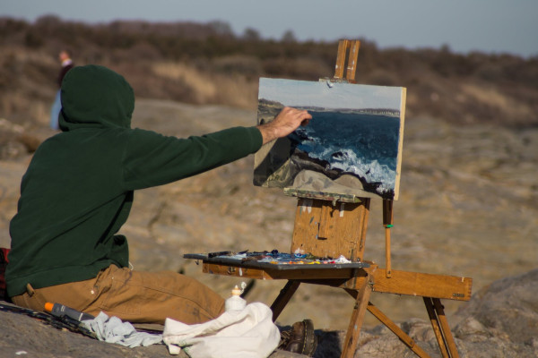 A color photography on an artist in a green hoodie painting a coastal scene on an easel set up on a rocky shoreline. The rocks have a tan color, the easel is unstinted wood, and the painter is wearing tan/brown pants.
