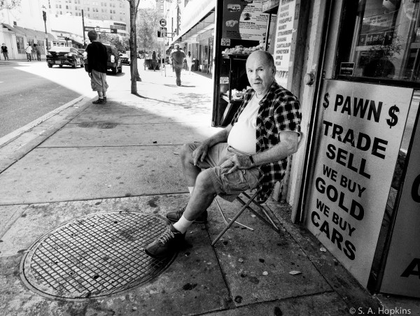 Black and white photo of a man sitting on a city sidewalk in front a pawn shop with a sign reading: trade, sell, we buy gold. 