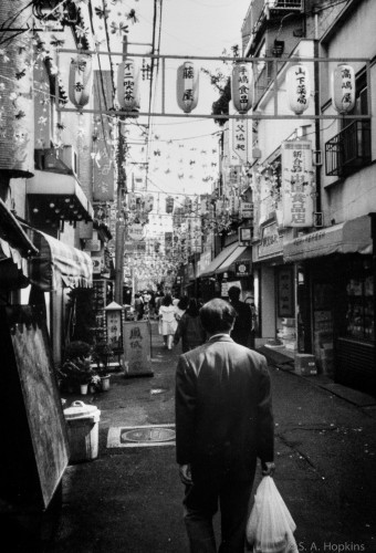The back of a man wearing a dark suit walking down a narrow street in Japan. There are various decorations adorning the buildings above. Black and white photo. 