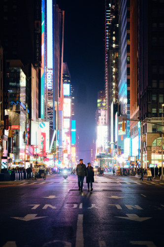A couple walks down an empty avenue in Times Square, the bright lights from the billboards on each side illuminate the black asphalt around them.

The couple is at the center of the frame, a bit lower than the middle. The person on the left is taller. They're wearing a baseball cap, dark green jacket, blue jeans and black leather shoes. The person on the right is shorter. They're wearing a black beanie, a black fur lined coat, blue jeans, and tan shoes.

The lights behind them are divided down the middle of the street on the left and right side of the frame, making a stark difference of scale, like the couple is at the bottom of a ravine of light.