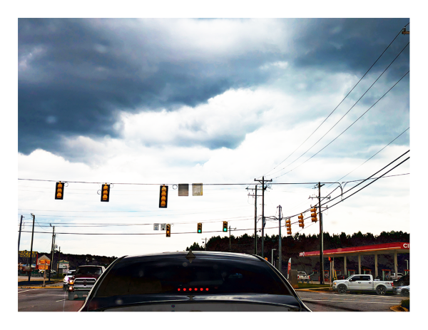 through the windshield, stopped at an semi-rural intersection in late afternoon under a stormy sky. we see signal lights, power lines, a circle k ahead on the right. two pick-up trucks. in the foreground, the trunk and rear windshield of a black car. six small, red brake lights are centered at the bottom.