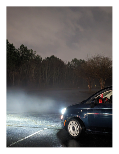 night, an empty, rain-slicked parking lot. a midnight blue economy car is parked in the foreground, its headlights illuminating fog with trees in the background, under a light, cloudy sky.