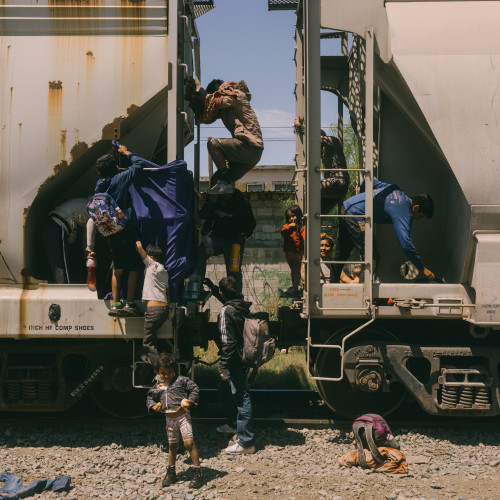 A pair of families photographed in the process of disembarking from their hiding places aboard La Bestia. A young child stands unsteadily on the ground while other family members climb from or cling to the sides and ladder of the train. One backpack has already been dropped to the gravel below the tracks.