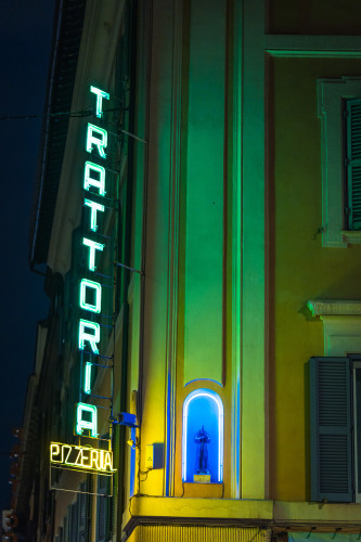 A building in Rome at night with a neon sign saying "Trattoria" and "Pizzeria". Next to it is a small figure in the building, supposedly a saint, lit up by blue neon light. 