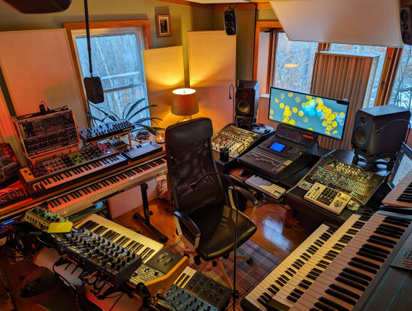 A music studio FULL of synthesizers and recording gear overlooking a wooded mountainside.