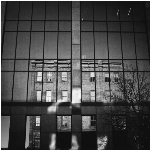 The reflection of a building can be seen in the windows of a large, tall glass sided office building in this black and white photo.