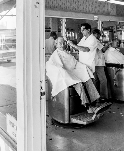 Black and white photo showing a man seated in a barber chair while the barber, wearing a guayabera, gives him a trim. The scene is photographed through the entryway. 