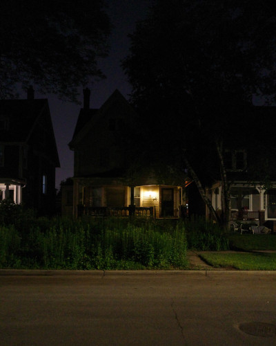 Color photograph of a house at night with a large porch lit brightly by a porchlight and a front yard filled with very tall plants